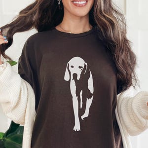 Puede incluir: Camiseta marrón oscuro con un gráfico blanco de un perro. El perro se representa en un estilo minimalista. La camiseta es de estilo casual, adecuada para el uso diario.