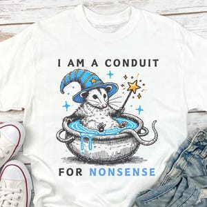 May include: White t-shirt with a graphic of a possum wearing a wizard hat and holding a wand. The possum is in a cauldron with blue liquid. The text "I am a conduit for nonsense" is printed on the shirt.