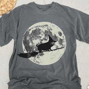 May include: Dark grey t-shirt with a black pug wearing a witch's hat, riding a broomstick in front of a full moon. The design is a whimsical illustration, suitable for Halloween or dog enthusiasts.