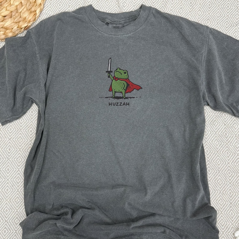 May include: Gray t-shirt featuring a cartoon frog wearing a red cape and holding a sword. The frog is standing on a dotted line, with the word "Huzzah" printed below. The shirt has a crew neck and short sleeves.