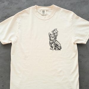 May include: Off-white t-shirt with a black and white illustration of a corgi in knight's armour. The corgi is depicted with a helmet, armour, and a sword. The shirt has a crew neck and short sleeves.