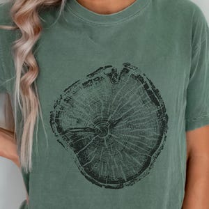 May include: Green t-shirt with a black tree ring design. The short-sleeved top is made of a soft material. The graphic is a detailed illustration of a tree's cross-section, showing growth rings.