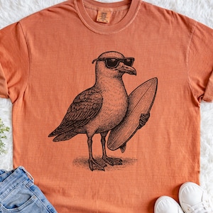 K&ouml;nnte beinhalten: Ein kurz&auml;rmeliges T-Shirt in gebranntem Orange mit einer schwarzen Grafik im Tintenstil, die eine M&ouml;we mit Sonnenbrille zeigt, die ein Surfbrett unter dem Fl&uuml;gel h&auml;lt. Das Shirt hat einen Rundhalsausschnitt und ein sichtbares Markenetikett.