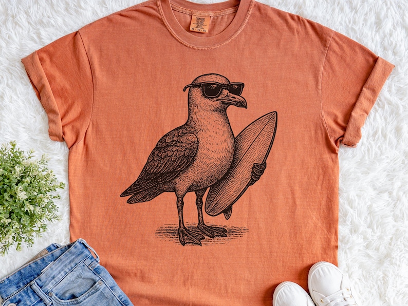 K&ouml;nnte beinhalten: Ein kurz&auml;rmeliges T-Shirt in gebranntem Orange mit einer schwarzen Grafik im Tintenstil, die eine M&ouml;we mit Sonnenbrille zeigt, die ein Surfbrett unter dem Fl&uuml;gel h&auml;lt. Das Shirt hat einen Rundhalsausschnitt und ein sichtbares Markenetikett.