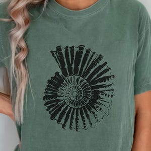 Może przedstawiać: Szałwiowa zielona koszulka z dużym, czarnym wzorem skamieniałości amonitu. Wzór, skamieniałość w kształcie spirali, jest umieszczony centralnie. Koszulka ma luźny krój i krótkie rękawy, idealna do noszenia na co dzień. Kolor jest stonowany i ziemisty.