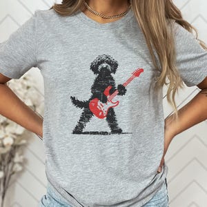 May include: A gray t-shirt featuring a black dog playing a red electric guitar. The dog is depicted in a distressed, textured style. The guitar has white accents. The shirt is a casual, everyday wear item.