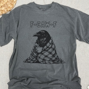 May include: Gray t-shirt with the text "F-CAW-F" above a black illustration of a raven wrapped in a plaid blanket, holding a steaming mug. The shirt is a casual wear item.