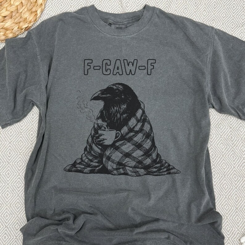 May include: Gray t-shirt with the text "F-CAW-F" above a black illustration of a raven wrapped in a plaid blanket, holding a steaming mug. The shirt is a casual wear item.