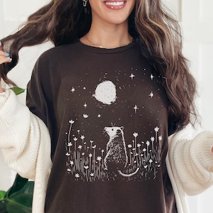 Pode incluir: Camiseta marrom escuro com uma ilustração branca de um gambá olhando para a lua e as estrelas. O design inclui flores brancas. A camiseta faz parte de uma coleção de roupas com tema da natureza.