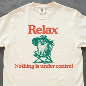May include: Off-white t-shirt with the word "Relax" in orange above a green illustration of a dog wearing sunglasses and a hat, sitting in a beach chair. Below the illustration, the text reads "Nothing is under control."