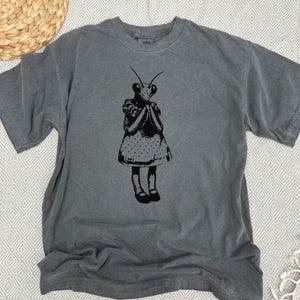 Puede incluir: Camiseta gris con una ilustraci&oacute;n negra de una mantis religiosa con un vestido de lunares y zapatos. El insecto se muestra con las manos juntas. La camiseta tiene cuello redondo y mangas cortas.