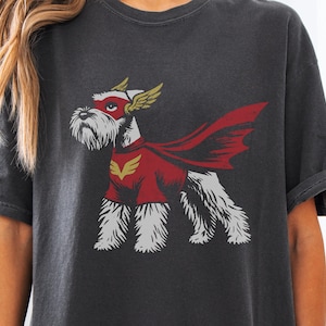 May include: Dark gray t-shirt featuring a cartoon Schnauzer superhero. The dog wears a red cape and mask, with gold wing accents. The design is printed on a soft, comfortable tee.