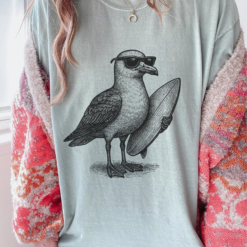 K&ouml;nnte beinhalten: Ein helles, salbeigr&uuml;nes T-Shirt mit Rundhalsausschnitt und einer schwarzen Grafik einer M&ouml;we, die eine Sonnenbrille tr&auml;gt und ein Surfbrett unter dem Fl&uuml;gel h&auml;lt. Das Shirt wird unter einer flauschigen Strickjacke mit orange-rosa &Auml;rmeln getragen.