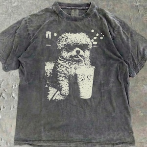 Peut inclure: Un t-shirt gris anthracite avec un motif blanc d'un chien portant des lunettes de soleil et buvant dans un gobelet avec une paille. Le t-shirt a un aspect vintage et délavé, avec un col rond.