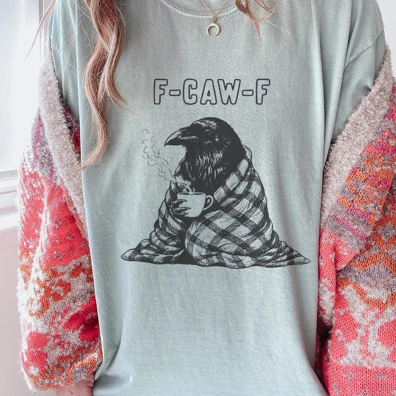 May include: A light blue t-shirt with the text "F-CAW-F" above a graphic of a raven wrapped in a plaid blanket, holding a mug. The raven is depicted with steam rising from the mug.