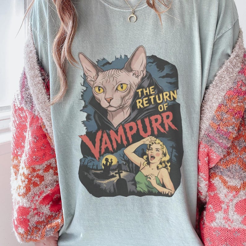 Peut inclure: T-shirt bleu clair avec un motif graphique. Le motif comprend le texte "THE RETURN OF VAMPURR", un chat Sphynx en cape de vampire, une femme qui crie et une sc&egrave;ne de cimeti&egrave;re.