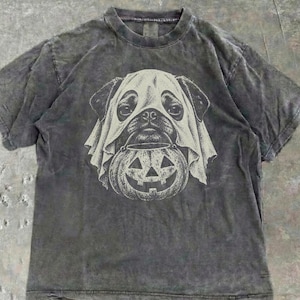 May include: A charcoal grey, acid-washed short-sleeve t-shirt featuring a graphic print of a pug wearing a white ghost sheet over its head. The dog is holding a carved jack-o'-lantern pumpkin bucket in its mouth, rendered in a detailed line art style.