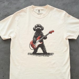 Peut inclure: T-shirt crème avec une illustration noire d'un chien jouant d'une guitare électrique rouge. Le chien est debout, tenant la guitare avec des détails blancs et rouges. Le t-shirt a un col rond et des manches courtes.