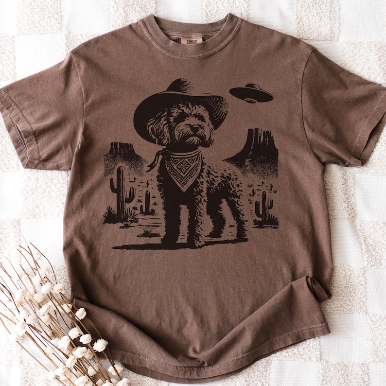 Pu&ograve; includere: T-shirt marrone con grafica nera di un cane con cappello da cowboy e bandana, ambientato in un paesaggio desertico con cactus e un UFO. La maglietta ha un aspetto vintage e usurato.