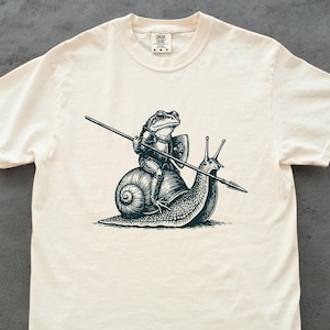 Könnte beinhalten: Cremefarbenes T-Shirt mit einer Schwarz-Weiß-Illustration eines Frosches in Rüstung, der eine Schnecke reitet und einen Speer hält. Die Schnecke hält ebenfalls einen Speer. Das Design ist auf dem Shirt zentriert.