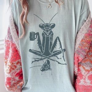 May include: A light blue t-shirt featuring a stylized illustration of a praying mantis holding a mug. The mantis is seated on crossed branches. The shirt has a vintage, distressed look.