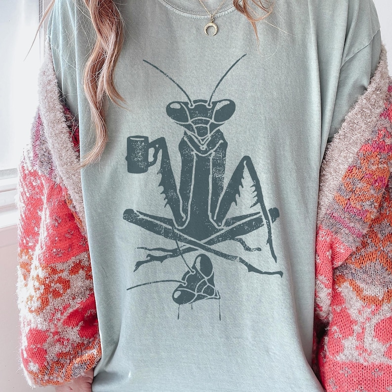 May include: A light blue t-shirt featuring a stylized illustration of a praying mantis holding a mug. The mantis is seated on crossed branches. The shirt has a vintage, distressed look.