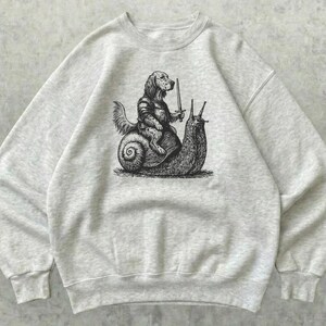 Op de afbeelding: Een lichtgrijs gemêleerd sweatshirt met ronde hals en een zwarte grafische print in inktstijl van een hond in middeleeuws harnas die een zwaard vasthoudt terwijl hij op de rug van een grote slak rijdt. Het kledingstuk heeft lange mouwen.