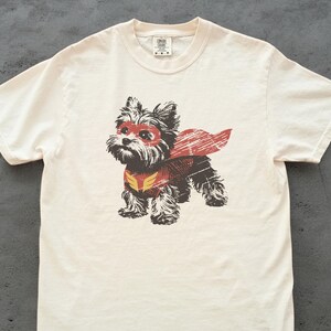 Pode incluir: Camiseta creme com um desenho de um cão vestido de super-herói. O cão usa uma máscara e capa vermelhas, com um emblema amarelo e vermelho no peito. Gola redonda e mangas curtas.