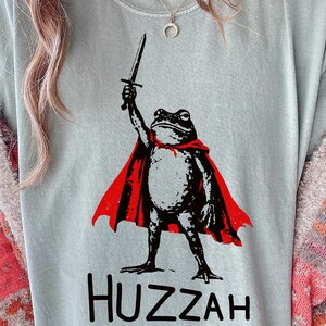 Op de afbeelding: Lichtblauw T-shirt met een zwart-witte illustratie van een kikker met een rode cape en een zwaard. Het woord "Huzzah" staat eronder in het zwart gedrukt.