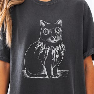 May include: Dark gray t-shirt featuring a detailed white illustration of a cat wearing a fish necklace. The cat is sitting upright, with large eyes and a detailed fur pattern. The shirt has rolled-up sleeves.