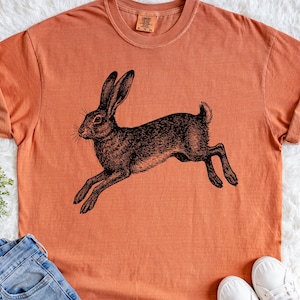 May include: An orange-colored t-shirt featuring a detailed black illustration of a running hare. The hare is depicted in a vintage, etched style, with long ears and outstretched legs. The shirt is casual and appears to be made of soft material.
