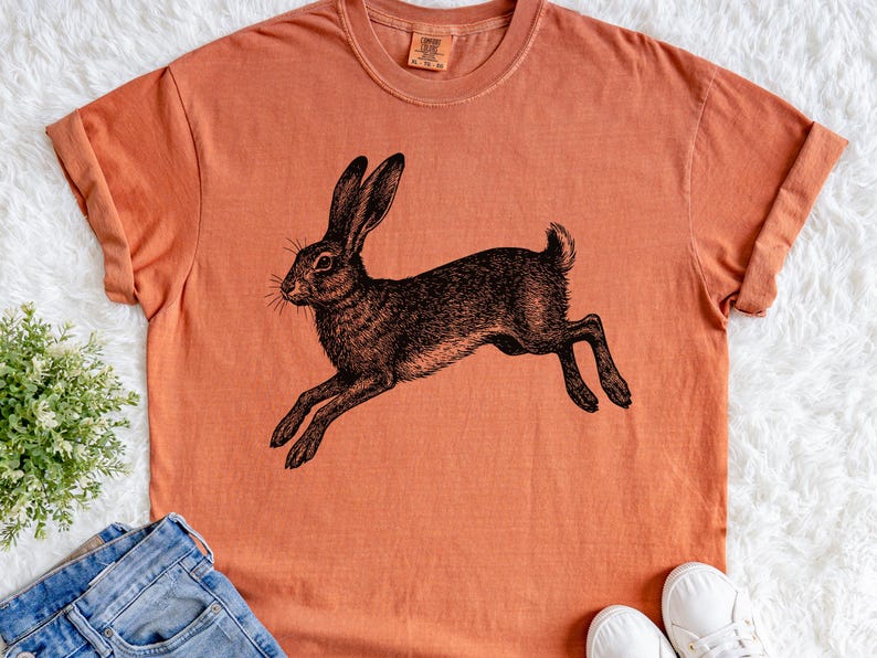 May include: An orange-colored t-shirt featuring a detailed black illustration of a running hare. The hare is depicted in a vintage, etched style, with long ears and outstretched legs. The shirt is casual and appears to be made of soft material.