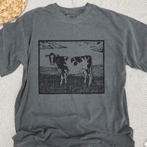 May include: Dark gray t-shirt featuring a black and white cow illustration within a rectangular frame. The cow stands in a field under a cloudy sky. The shirt has short sleeves and a crew neck.