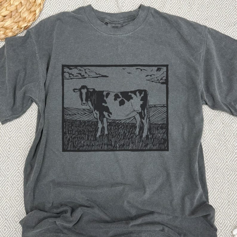 May include: Dark gray t-shirt featuring a black and white cow illustration within a rectangular frame. The cow stands in a field under a cloudy sky. The shirt has short sleeves and a crew neck.