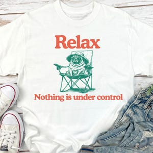 May include: White t-shirt featuring a pug wearing sunglasses and a hat, seated in a green folding chair. The text "Relax" and "Nothing is under control" are in orange.