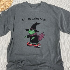 以下が含まれることがあります： 「Off to write code」の文字が入ったグレーのTシャツ。魔女の帽子、スカーフ、マントを着た緑色のカエルが赤いホバーボードに乗っているイラスト。カエルは黒いアクセントで描かれています。