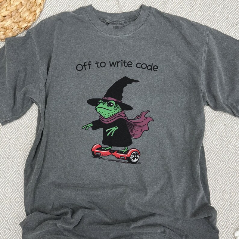 以下が含まれることがあります： 「Off to write code」の文字が入ったグレーのTシャツ。魔女の帽子、スカーフ、マントを着た緑色のカエルが赤いホバーボードに乗っているイラスト。カエルは黒いアクセントで描かれています。