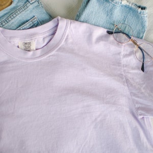 May include: A light purple Comfort Colors t-shirt with a crew neck, laid on top of light blue denim jeans. A pair of round, gold-rimmed glasses rests on the jeans. The t-shirt has a tag that reads "Comfort Colors".