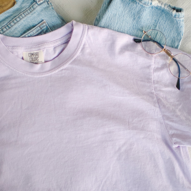 May include: A light purple Comfort Colors t-shirt with a crew neck, laid on top of light blue denim jeans. A pair of round, gold-rimmed glasses rests on the jeans. The t-shirt has a tag that reads "Comfort Colors".