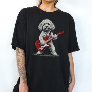 May include: A black t-shirt featuring a graphic illustration of a fluffy, light-coloured dog standing on its hind legs while playing a bright red electric guitar. The dog has a textured, sketch-like appearance, and the guitar has white accents.