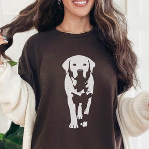 May include: A dark brown t-shirt featuring a white graphic of a Labrador Retriever dog. The dog is depicted in a walking pose. The person is wearing a white cardigan over the t-shirt.