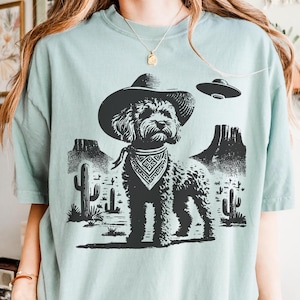Puede incluir: Camiseta azul claro con un gráfico negro de un perro con sombrero de vaquero y pañuelo, sobre un paisaje desértico con cactus y montañas. Un OVNI flota sobre el perro.