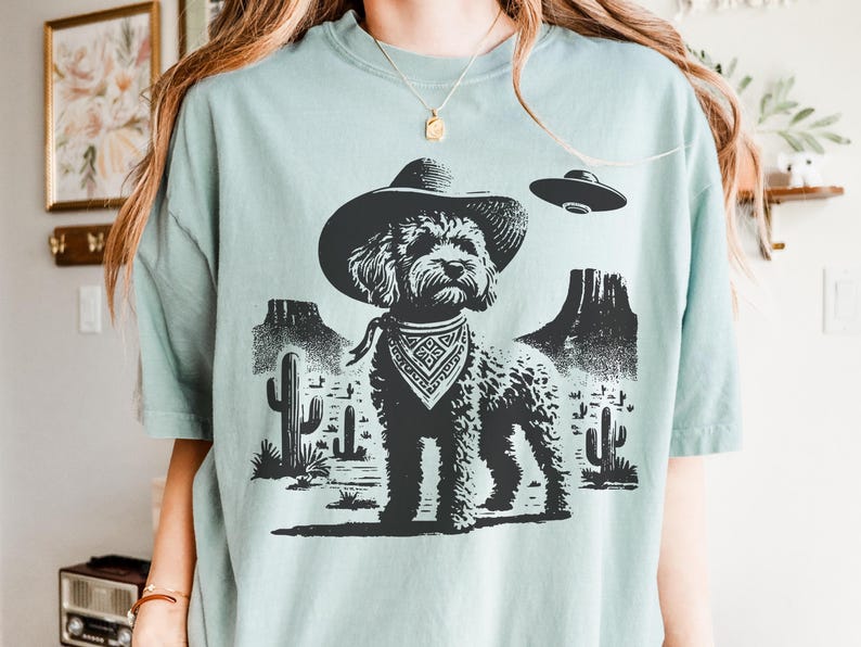 Pu&ograve; includere: Maglietta azzurra con grafica nera di un cane con cappello da cowboy e bandana, ambientata in un paesaggio desertico con cactus e montagne. Un UFO fluttua sopra il cane.