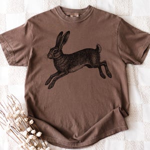 May include: Brown t-shirt featuring a detailed black illustration of a running rabbit. The shirt has a classic crew neck and short sleeves. The rabbit illustration is centered on the front of the shirt, creating a vintage aesthetic.