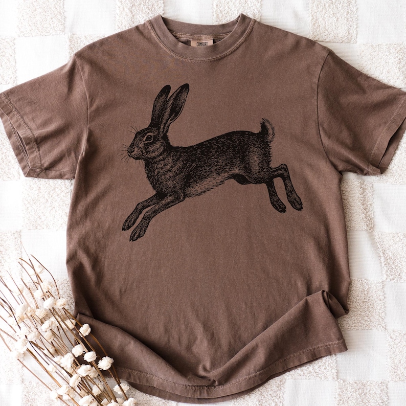 May include: Brown t-shirt featuring a detailed black illustration of a running rabbit. The shirt has a classic crew neck and short sleeves. The rabbit illustration is centered on the front of the shirt, creating a vintage aesthetic.