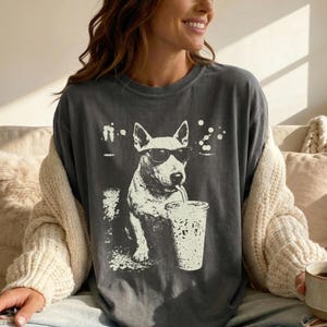 Peut inclure: T-shirt gris foncé avec un motif blanc d'un chien portant des lunettes de soleil et buvant dans un gobelet. Le chien est au centre, avec une ambiance décontractée. Le t-shirt est un vêtement confortable pour tous les jours.