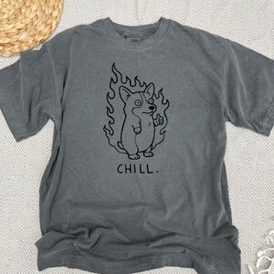 May include: Gray t-shirt featuring a black graphic of a corgi dog surrounded by flames, giving a thumbs-up. The word "CHILL" is printed below the dog. The shirt has short sleeves and a crew neck.