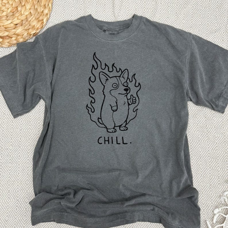 May include: Gray t-shirt featuring a black graphic of a corgi dog surrounded by flames, giving a thumbs-up. The word "CHILL" is printed below the dog. The shirt has short sleeves and a crew neck.