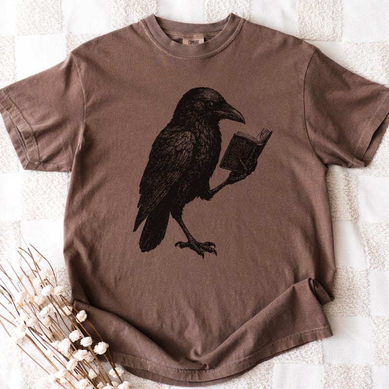 May include: A brown t-shirt featuring a black illustration of a raven reading a book. The raven is depicted in a detailed, vintage style. The t-shirt has a crew neck and short sleeves, with a slightly faded appearance.