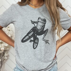 Pu&ograve; includere: Una maglietta grigia con una grafica nera di una rana in bicicletta. La rana &egrave; raffigurata in uno stile illustrativo dettagliato, con le zampe e le braccia posizionate come se stesse pedalando e guidando. Il colore unito della maglietta offre uno sfondo neutro per il design stravagante.